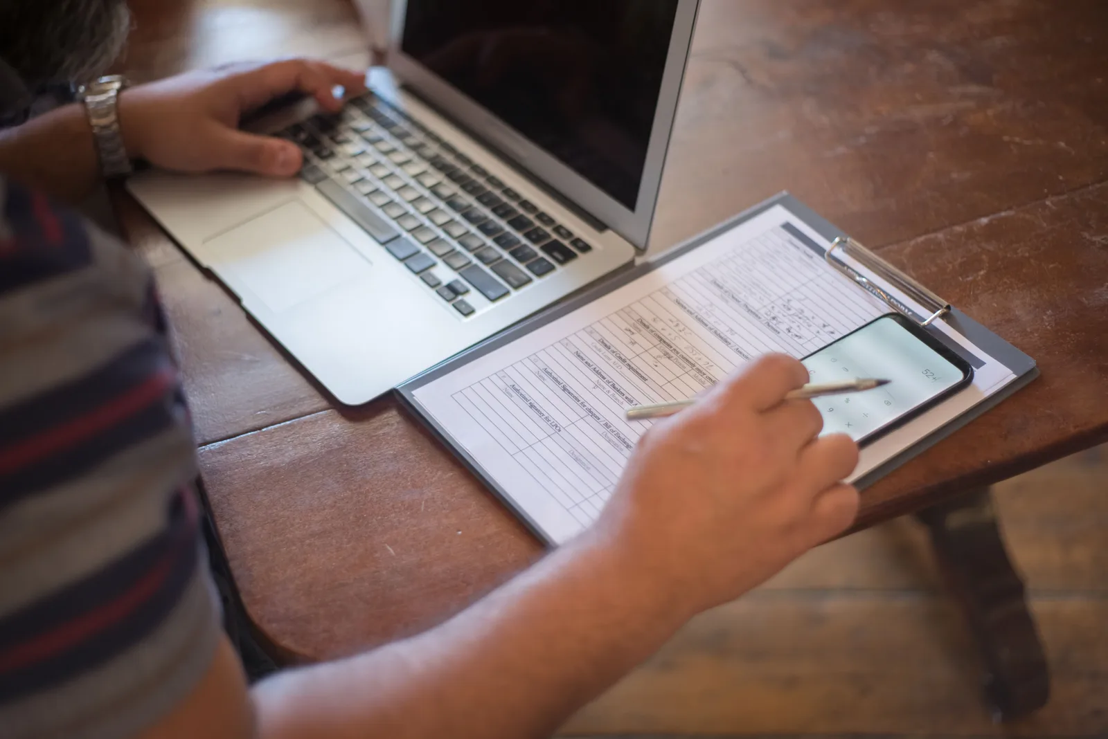 Close-up of a person using a laptop and reviewing documents with a smartphone on a wooden table. Beyond handyman licensing requirements: compliance details