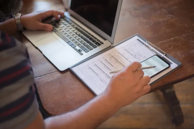 Close-up of a person using a laptop and reviewing documents with a smartphone on a wooden table. Beyond handyman licensing requirements: compliance details